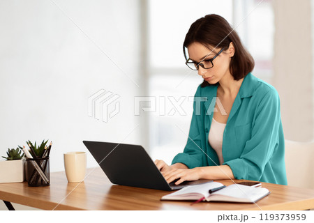 Home Office. Portrait of young female manager wearing specs and brown shirt using laptop, working remotely. Copy space 119373959