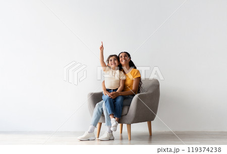 Check This. Happy Young Mother And Little Daughter Sitting In Armchair And Pointing Up, Cheerful Middle Eastern Mom And Female Child Demonstrating Copy Space Above Their Heads While Resting In Chair Check This. Happy Young Mother And Little Daughter Sitting In Armchair And Pointing Up, Cheerful Middle Eastern Mom And Female Child Demonstrating Copy Space Above Their Heads While Resting In Chair 119374238