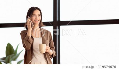 Communication Concept. Young manager talking on mobile phone, standing next to window, holding coffee cup 119374676