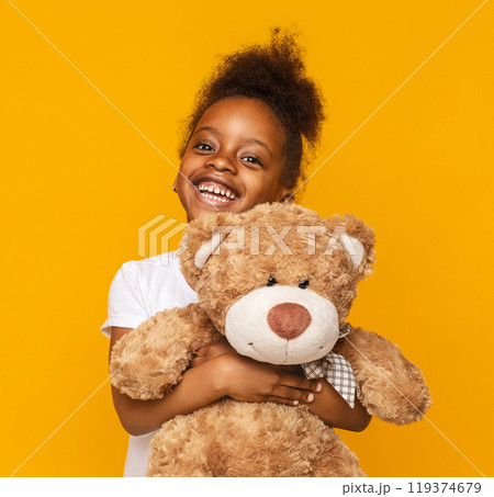 My favorite toy. Cute little african girl hugging her teddy bear and smiling, orange studio background 119374679