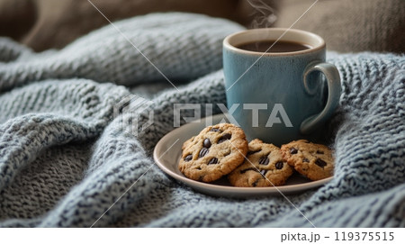 A steaming cup of coffee sits beside chocolate chip cookies on a plate, surrounded by a warm, textured blanket. 119375515