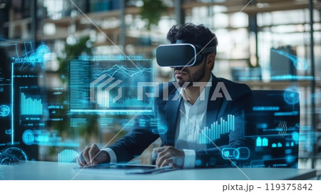 A man wearing a virtual reality headset sits at a desk in an office. The headset displays various graphs and data visualizations 119375842