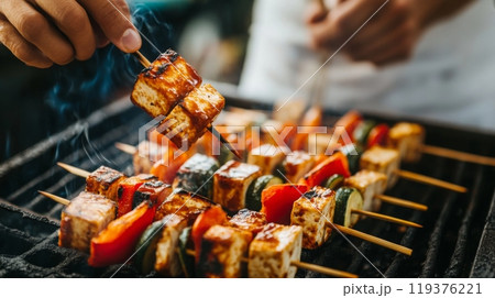 Skewers of marinated tofu and colorful vegetables are grilled, emitting savory smoke at an outdoor barbecue gathering. 119376221