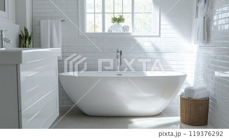 A stylish bathroom features a freestanding white tub illuminated by natural light. The space is designed with a minimalist aesthetic, showcasing white tiles, sleek cabinetry, and decorative plants. 119376292