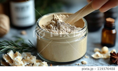 A person stirs a jar of fine natural powder with a wooden spoon, accompanied by herbal elements and essential oil bottles on a rustic countertop. 119376488