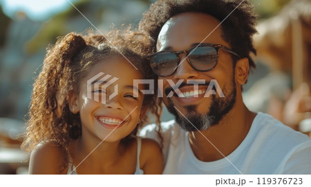 A father and his daughter share a bright day at the beach, smiling widely in the sun. Their bond is evident as they enjoy each other's company in a warm, relaxed atmosphere. 119376723