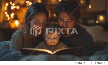 A couple sits comfortably on a bed, reading a bedtime story to their smiling toddler. The room is softly illuminated by warm lights, creating a serene atmosphere. 119376853