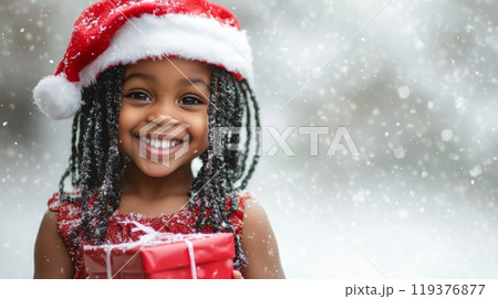 A smiling child wearing a festive Santa hat stands in the snow, holding a beautifully wrapped Christmas gift. The joy of the season is evident as snowflakes gently fall around her. 119376877