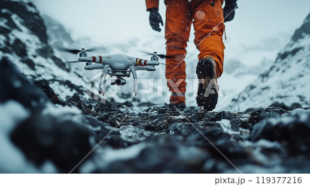A person dressed in bright orange walks on rocky ground covered with snow, controlling a drone hovering nearby in the chilly mountain setting. 119377216