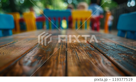 A close-up view of a weathered wooden table with blurred colorful chairs in the background. 119377519