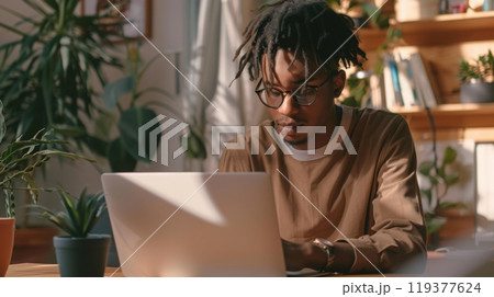 A young man with dreadlocks concentrates on his laptop in a bright home office filled with plants. 119377624