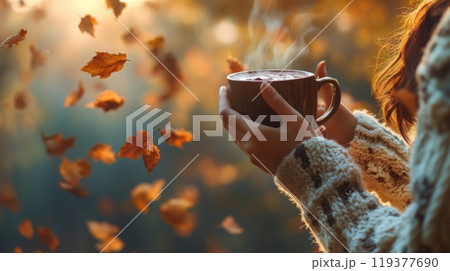 A person holds a steaming cup of a warm drink as colorful autumn leaves drift gently through the air. The soft sunlight illuminates the scene, creating a cozy and tranquil atmosphere. 119377690