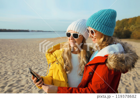 Happy young women friends with smartphone together on beach, smiling girlfriends using phone on sea Happy young women friends with smartphone together on beach, smiling girlfriends using phone on sea 119378682