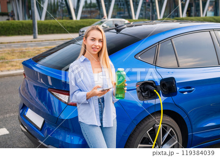 A woman stands by her electric car, charging while holding a green bottle and using her phone to pay. Green energy concept 119384039