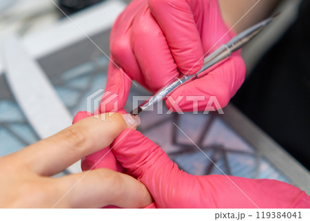 Applying a base coat and gel polish during a manicure in a nail salon by nail master in rubber gloves Applying a base coat and gel polish during a manicure in a nail salon by nail master in rubber gloves 119384041