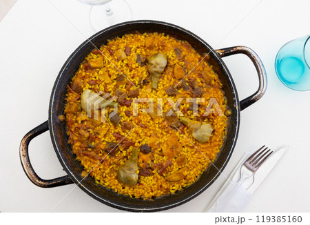 Closeup of rich flavorful paella in traditional pan, generously filled with meat, bacon pieces and artichokes. Culinary traditions of Spain Closeup of rich flavorful paella in traditional pan, generously filled with meat, bacon pieces and artichokes. Culinary traditions of Spain 119385160