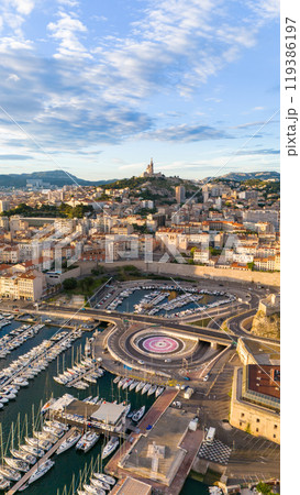 Aerial view of Marseille, France, featuring the vibrant port with yachts, the historic Fort Saint-Nicolas, and the iconic Notre-Dame de la Garde basilica atop the hill. 119386197