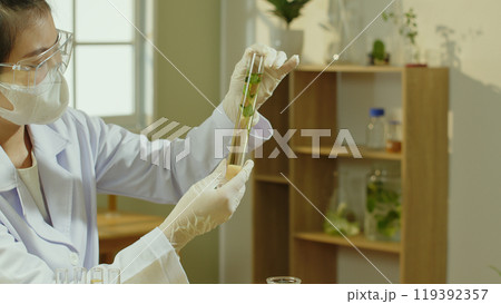 This high-quality photo of a laboratory space features a scientist holding a tube containing herbal solution inside for observation, behind which is a wooden shelf displaying other tools and herbs. This high-quality photo of a laboratory space features a scientist holding a tube containing herbal solution inside for observation, behind which is a wooden shelf displaying other tools and herbs. 119392357