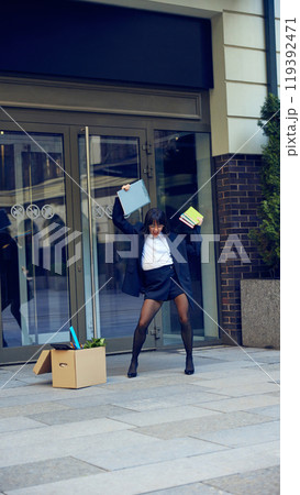 Happy young woman cheerfully dancing after quitting hated job, walking out the office building with box filled with her stuffs Happy young woman cheerfully dancing after quitting hated job, walking out the office building with box filled with her stuffs 119392471