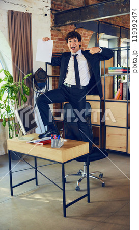 Happy emotional young man in formal wear standing in office, pointing at document, expressing happiness about getting promotion Happy emotional young man in formal wear standing in office, pointing at document, expressing happiness about getting promotion 119392474
