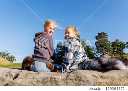 Back view two cute little girls friends wearing casual hoodies plaid jackets and attached fox fur tails enjoy sit on rock on warm sunny day outdoors. Clear blue sky trees woods background 119395372