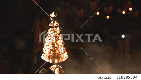 Varanasi, India. Man makes movements with a candle holder with lit candles on the Ganga Maha Aarti ceremony begins. Brahman priest wearing red and yellow dress doing traditional ganga aarti at ghat 119395394
