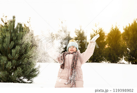 Happy child portrait. Excited kid playing in winter mountains forest. Funny little girl throwing snow, having fun. Snowy evergreen trees around ski resort. Christmas holiday vibes on sunny day outdoor 119398902