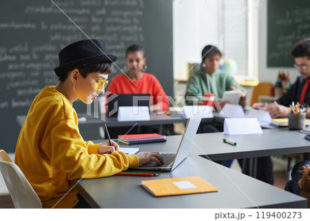 Side view portrait of teenage girl sitting at table and using computer during lesson in school classroom, copy space Side view portrait of teenage girl sitting at table and using computer during lesson in school classroom, copy space 119400273
