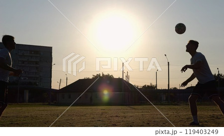 Silhouette of professional footballers kicking soccer ball with their heads on stadium at sunset. Two sportsmen showing tricks with ball while passing it to each other at field. Freestyle football 119403249