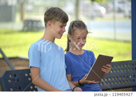 Two happy children, girl and boy looking at screen of digital tablet, reading, studying or gaming sitting on bench outdoors on summer sunny day Two happy children, girl and boy looking at screen of digital tablet, reading, studying or gaming sitting on bench outdoors on summer sunny day 119405427