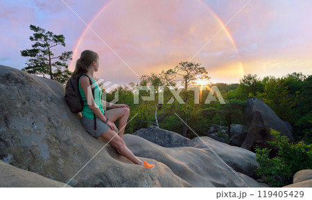 Sportive woman sitting alone taking a break on hillside trail. Female hiker enjoying view of evening nature from rocky cliff on wilderness path. Active lifestyle concept 119405429