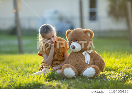 Sad lonely child girl spending time together with her teddy bear friend outdoors on sunny backyard. Loneliness at preteen age Sad lonely child girl spending time together with her teddy bear friend outdoors on sunny backyard. Loneliness at preteen age 119405433