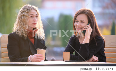 Female friends spending time at street cafe outdoors while one girl is talking happily on mobile phone and another waiting frustrated at offensive behaviour of her mate. Friendship problems concept Female friends spending time at street cafe outdoors while one girl is talking happily on mobile phone and another waiting frustrated at offensive behaviour of her mate. Friendship problems concept 119405474