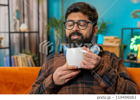 Attractive smiling Indian young man drinking a cup of coffee or herbal tea at home couch in morning Attractive smiling Indian young man drinking a cup of coffee or herbal tea at home couch in morning 119406061