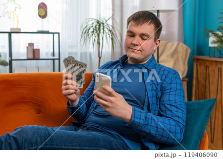 Successful rich man holding smartphone and counting dollar bill money cash sitting on home sofa Successful rich man holding smartphone and counting dollar bill money cash sitting on home sofa 119406090