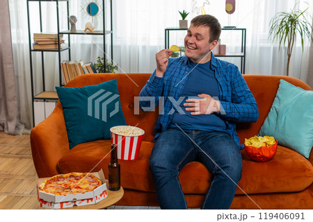 Happy fat man sitting on sofa eating junk food chips pizza popcorn and beer, watching TV film game Happy fat man sitting on sofa eating junk food chips pizza popcorn and beer, watching TV film game 119406091