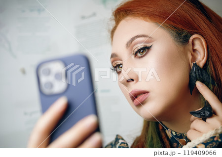 Red-haired woman takes selfie on front camera of modern smartphone. Female blogger films herself for fashion and makeup blog. Cute girl points her finger at her butterfly earrings. Gothic aesthetic. Red-haired woman takes selfie on front camera of modern smartphone. Female blogger films herself for fashion and makeup blog. Cute girl points her finger at her butterfly earrings. Gothic aesthetic. 119409066