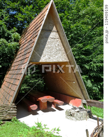 Triangular wooden hut for a picnic with a wooden tile roof. Red benches and table, barbecue hearth 119409199