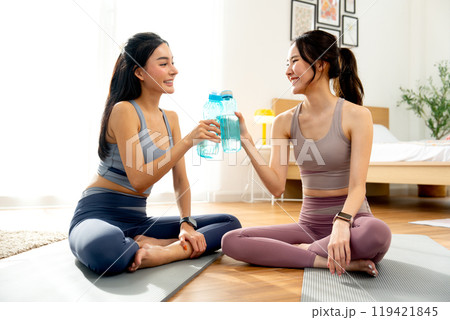 Two Asian women with exercise clothes sit on yoga mat and hold bottle of water then cheers together and relax after workout in the house. Two Asian women with exercise clothes sit on yoga mat and hold bottle of water then cheers together and relax after workout in the house. 119421845