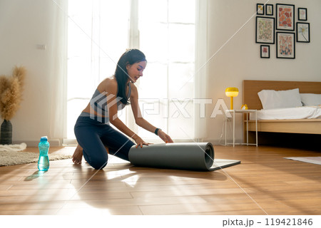 Side view pretty Asian woman with exercise clothes lay out a yoga mat to prepare for exercise in front of warm light throgh glass door. 119421846