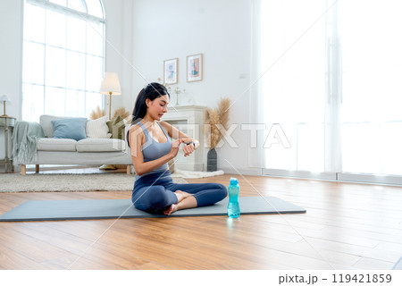 Pretty Asian woman with exercise clothes sit on mat in room with look at wristwatch during yoga exercise with happiness. Pretty Asian woman with exercise clothes sit on mat in room with look at wristwatch during yoga exercise with happiness. 119421859