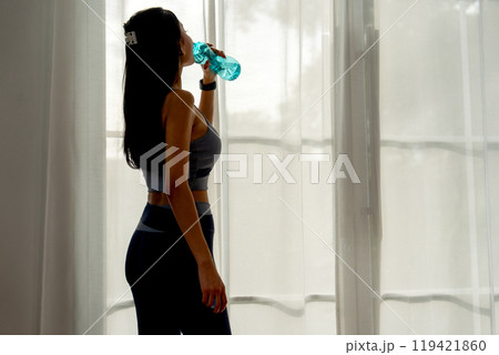 Back of pretty Asian woman with exercise clothes drink water from bottle also stand in front of curtain and glass door with happiness. 119421860