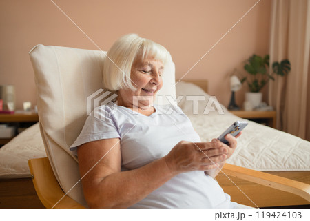 Caucasian 60s happy woman using cell phone relaxing at home. Smiling senior lady holding smartphone enjoying mobile shopping, checking social media messages, playing game or buying online. Caucasian 60s happy woman using cell phone relaxing at home. Smiling senior lady holding smartphone enjoying mobile shopping, checking social media messages, playing game or buying online. 119424103