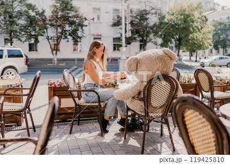 A woman sits cafe with a teddy bear next to her. The scene is set in a city with several chairs and tables around her. The woman is enjoying her time at the outdoor cafe. A woman sits cafe with a teddy bear next to her. The scene is set in a city with several chairs and tables around her. The woman is enjoying her time at the outdoor cafe. 119425810