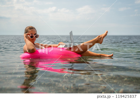 A woman is floating in a pink inflatable raft with a laptop in her hand. She is smiling and she is enjoying her time in the water. 119425837