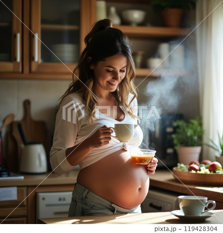 A smiling pregnant woman in white shirt holds a steaming cup while standing in a sunlit kitchen, emphasizing her baby bump 119428304