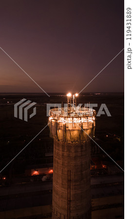 Vertical photo.Illuminated construction site at dusk.Dangerous work at night.Workers build a high chimney for a plant.Contractor repairs a chimney on a high-rise building.Worker in a hardhat on site 119431889