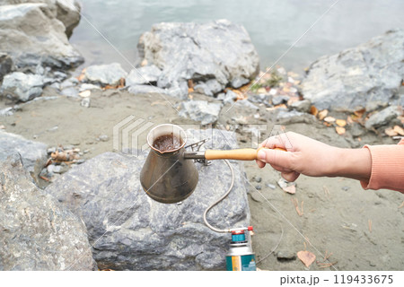 Outdoor coffee brewing by riverbank using traditional pot and camping stove 119433675