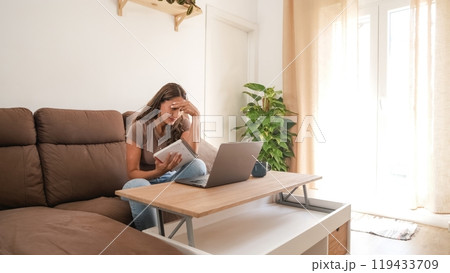 Stressed woman holding notebook while working on laptop at home 119433709