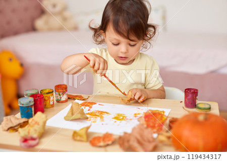 cute little child girl painting autumn leaves. baby girl drawing leaves in yellow and orange paint cute little child girl painting autumn leaves. baby girl drawing leaves in yellow and orange paint 119434317